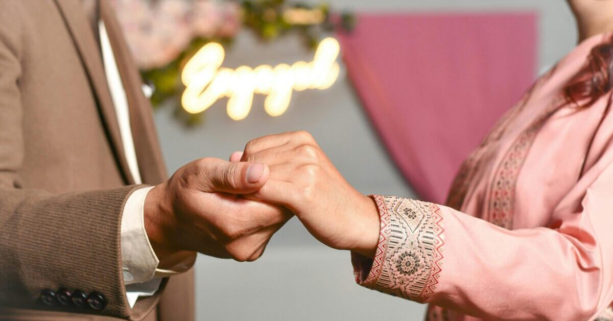 Close-up of couple's hands during an engagement, with 'Engaged' sign in background.