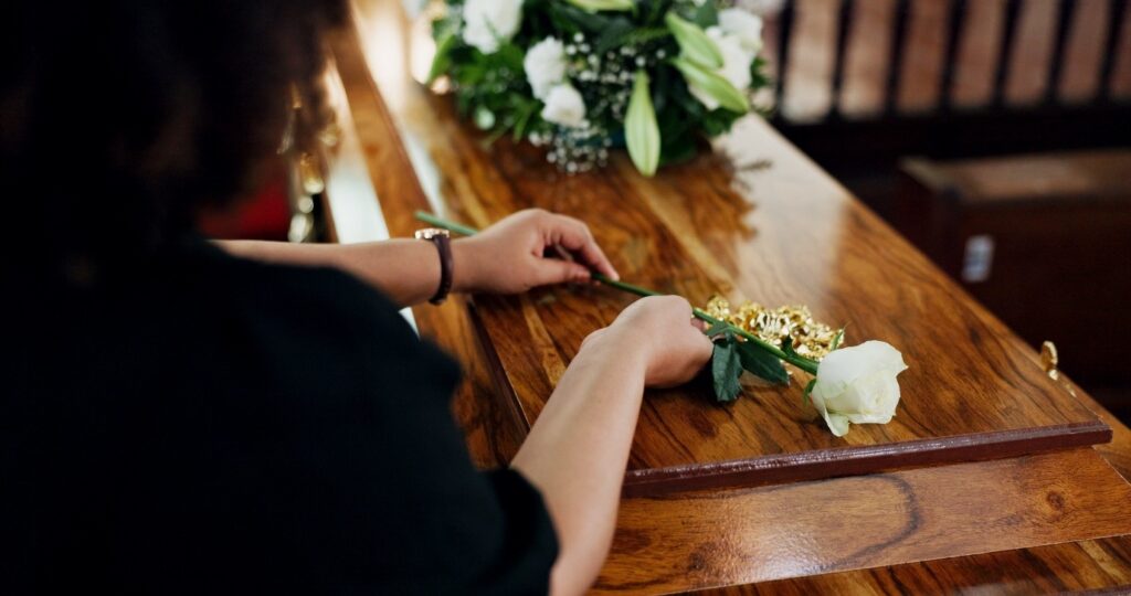 A woman, serving as a funeral celebrant, lays a flower on a wooden casket at a funeral service.