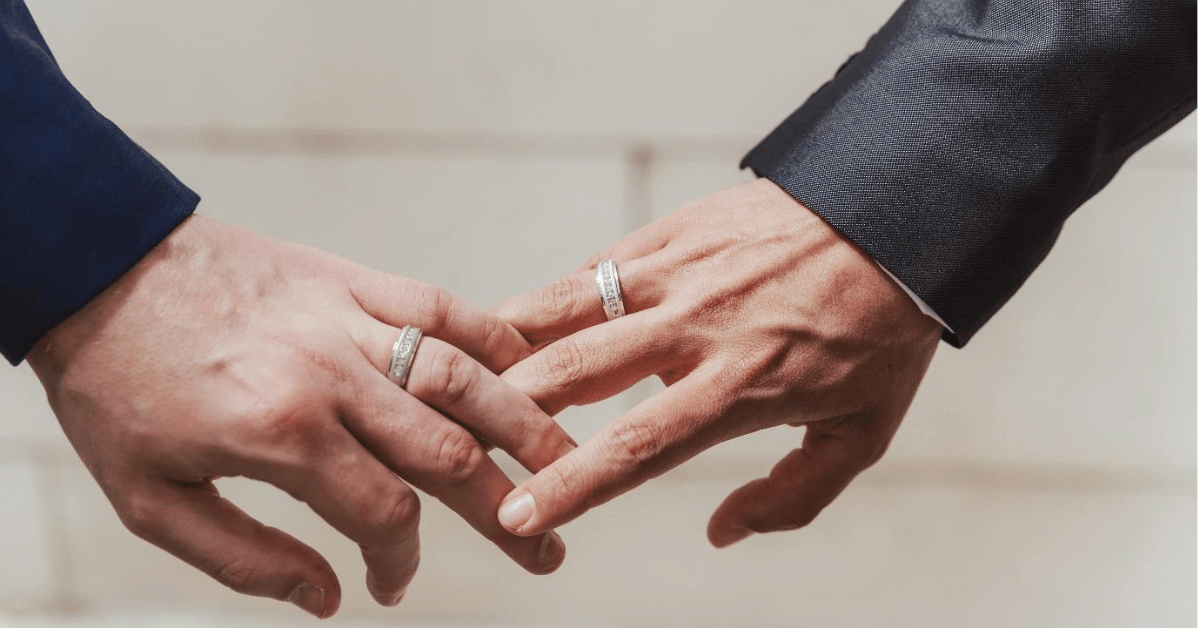 Two men in suits holding hands with wedding rings, marking their vow renewal ceremony led by a marriage celebrant.